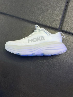 Vita Hoka Bond 8 skor - Snygga vita sneakers från Hoka med riktigt chunky och hög sula. Skorna har mesh-ovandel för bra andningsförmåga och Hoka-logga på sidan. Perfekta för dig som gillar sportig stil och vill ha maximal komfort när du är ute och går eller springer.