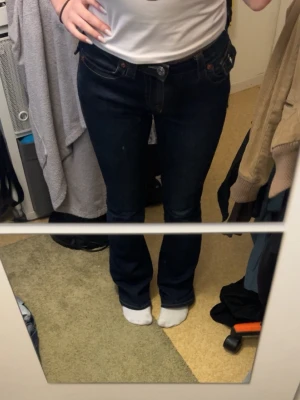 Mörkblå bootcut jeans True Religion - Säljer ett par mörkblå bootcut jeans från True Religion med klassiska kontrastsömmar och stora fickor bak med dekorativa knappar. Jeansen har låg midja och är tillverkade i kraftigt denimtyg. Perfekta för dig som gillar en tidlös och cool look.