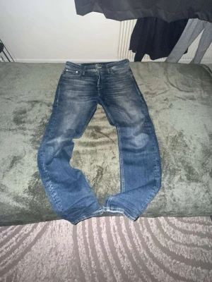 Blåa jeans från Jack & Jones  - Säljer ett par klassiska blå jeans med raka ben. Jeansen har fem fickor och normal midja. Perfekta till en avslappnad stil och passar till det mesta. Materialet är troligtvis bomull med strech så man är bekväm vart man än går med jeansen. Pass formen är regular inte för tajt och inte för baggy.