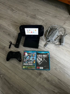 Nintendo Wii U med spel och tillbehör - Säljer en svart Nintendo Wii U med GamePad, Pro Controller, Wii Remote, laddare och två spel: Super Mario Bros. U och Call of Duty Black Ops II. Konsolen har en stilren design i plast och medföljande kablar för enkel uppkoppling. Perfekt för gamingkvällar med vänner.