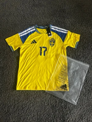 Sverige matchtröja hemma 2026 "17" - Gul svensk landslagströja storlek M från Adidas med blå detaljer, nummer 17 på bröstet och klassiskt landslagsemblem. Tröjan har korta ärmar, blå och vita ränder på axlarna och är i slim fit-modell. Perfekt för fotboll eller träning. Perfekt för att heja på sverige lite extra i vm. En replika
