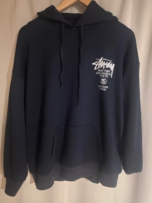 Mörkblå Stussy hoodie med tryck - Snygg mörkblå hoodie från Stussy med vit logga och tryck på både bröstet och ryggen. Klassisk känguruficka framtill och justerbar huva med dragsko. Perfekt för en streetwear-look och riktigt skön passform.