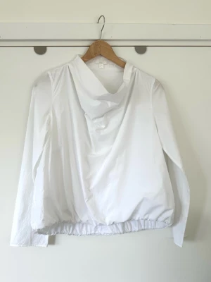 Vit långärmad blus med draperad krage - storlek S - Stilren vit oversize blus / topp från COS med en avslappnad passform. Storlek 34 / S, men passar även M om man vill att den ska sitta mer figurnära. Tidlös modell med vid draperad krage som kan bäras uppvikt eller nedvikt, vilket ger en mångsidig stil. Smockning nedtill för fin siluett. 100% bomull, så bra material som andas. Mycket gott skick, som ny!