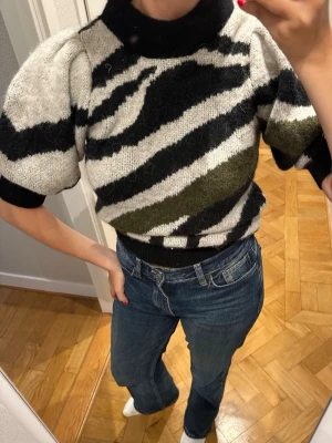Zebramönstrad stickad tröja - Säljer en kortärmad stickad tröja med zebramönster i svart, vitt och olivgrönt. Tröjan har puffiga ärmar, hög krage och ribbade muddar. Materialet känns mjukt och mysigt. Använt en gång men inga noppror inga defekter alls! 