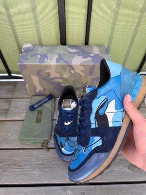 Valentino rockrunner camo sneakers - Unika sneakers från Valentino med blått camo-mönster, mörkblå mockadetaljer och vita inslag. Skorna har klassisk låg profil, blå snörning och gummisula. Kommer med originalkartong, extra skosnören och dustbag. Perfekta för dig som vill sticka ut med stil. 100% äkta 