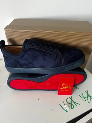 Mörkblå sneakers från CL och en grön plånbok - Snygga mörkblå sneakers från CL i mocka med klassisk röd sula. Skorna har lågt skaft, ton-i-ton snörning och diskret broderad logga på hälen. Perfekta för dig som gillar exklusiv och stilren design. Och en grön plånbok