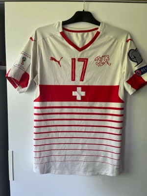 Schweiz matchtröja Puma nr 17 - Säljer en officiell Schweiz fotbollslandslagets matchtröja från Puma med nummer 17. Tröjan är vit med röda detaljer, ränder och Schweiz flagga på bröstet. Korta ärmar med tryck på båda, V-ringad krage och tillverkad i lätt funktionsmaterial.