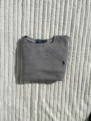 Grå långärmad tröja från Polo Ralph Lauren - Grå långärmad tröja från Polo Ralph Lauren i ribbad bomull med rund halsringning. Klassisk svart broderad logga på bröstet och breda muddar vid ärmslut. Perfekt för en clean och stilren look.