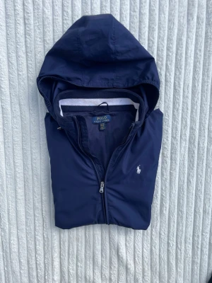 Marinblå vindjacka från Polo Ralph Lauren - Snygg marinblå vindjacka med huva från Polo Ralph Lauren. Jackan har dragkedja framtill, två fickor med zip och vit logga på bröstet. Elastiska muddar med vit detalj. Materialet är lätt och vattenavvisande, perfekt för blåsiga dagar.