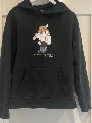 Svart hoodie Polo Ralph Lauren  - Svart hoodie från Polo Ralph Lauren med ikoniska Polo Bear-trycket på bröstet. Klassisk känguruficka framtill och huva. Tillverkad i mjukt bomullsmaterial som är skönt att ha på sig. Perfekt för dig som gillar streetwear och vill ha en stilren look.