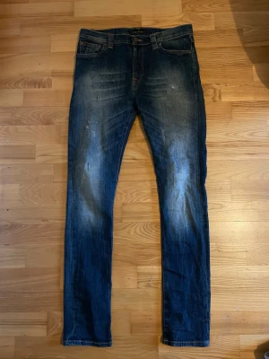 Blå slim fit jeans från Nudie Jeans - Säljer ett par blå slim fit jeans från Nudie Jeans med klassisk femficksmodell och snygga slitningar framtill. Jeansen har en något tvättad look och kontrastsömmar. Perfekta för dig som gillar en smalare passform och tidlös stil.
