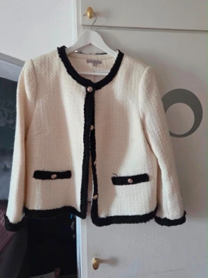 Vit bouclé kavaj med svarta detaljer H&M - Stilren vit bouclé kavaj från H&M med svarta kontrastkanter och dekorativa guldknappar. Rund halsringning, två fickor fram och rak passform. Perfekt för en chic och trendig look. Materialet är strukturerat och ger en exklusiv känsla.