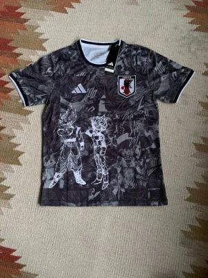 Adidas Japan x Dragon Ball Z tröja - Cool svartgrå sporttröja från Adidas med Japan-landslagets emblem och Dragon Ball Z-motiv på framsidan. Slim fit-modell med vita detaljer vid krage och ärmslut. Perfekt för dig som gillar anime och fotboll. Officiellt licensierad produkt. den är helt ny aldrig använd. jag är 182 och den sitter perfekt på mig. 