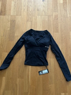 DFYNE Orgin Long Sleeve V-Neck Top - Säljer en DFYNE Orgin Long Sleeve V-Neck Top med inbyggd sport-bh i färgen ”Midnight Black” i st XS, oanvänd med lappen kvar. Original pris 56,95€ - ca 630kr.