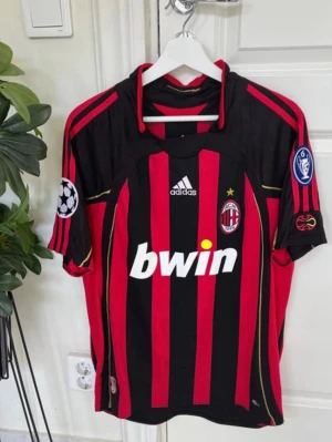 AC Milan matchtröja Kaka 22 Adidas - Snygg AC Milan matchtröja i svart och rött med guldiga detaljer, Adidas-logga och Bwin-tryck på bröstet. Champions League-märke på ärmen och Kaka 22 tryckt på ryggen. Klassisk fotbollströja i polyester, perfekt för fans och samlare.
