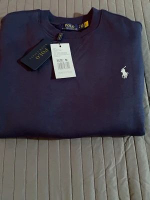Mörkblå sweatshirt från Polo Ralph Lauren - Klassisk mörkblå sweatshirt från Polo Ralph Lauren med rund halsringning och vit broderad logga på bröstet. Tröjan har långa ärmar och ribbade muddar vid ärmslut och nederkant. Perfekt för en stilren och avslappnad look.