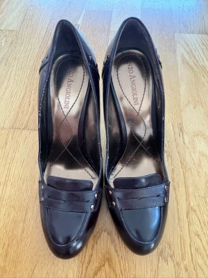 Bruna pumps från Enzo Angiolini strl 36 - Snygga mörkbruna pumps från Enzo Angiolini med hög klack och dekorativa nitar vid tån. Skorna har en klassisk penny loafer-inspirerad design och är tillverkade i blankt läder. Perfekta för dig som vill ha en stilren och elegant look. Klackhöjd 8-9cm. storlek 36. I fint skick