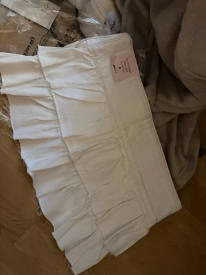 Vit volangkjol från Gina Tricot - Supersöt vit kortkjol från Gina Tricot med volangdetaljer. Kjolen har en hög midja och är tillverkad i ett mjukt bomullstyg. Perfekt för dig som vill ha en fräsch och trendig look med lite extra volym.