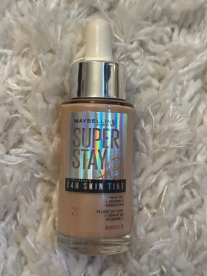 Maybelline Super Stay 24H Skin Tint - Foundation från Maybelline i nyansen 21, Super Stay 24H Skin Tint med vitamin C. Kommer i en glasflaska med pipett och har en beige ton. Perfekt för dig som vill ha en naturlig och fräsch bas med lång hållbarhet.