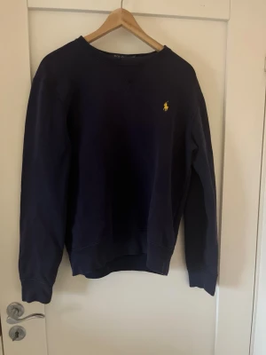 Mörkblå sweatshirt från Polo Ralph Lauren - Klassisk mörkblå sweatshirt från Polo Ralph Lauren med rund halsringning och gul broderad logga på bröstet. Tröjan har ribbade muddar vid ärmslut och nederkant. Perfekt för en avslappnad och stilren look.