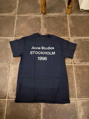 Mörkblå Acne Studios t-shirt - Snygg mörkblå t-shirt från Acne Studios med vit text 'STOCKHOLM 1996' på bröstet. Klassisk passform med rund hals och korta ärmar. Perfekt för dig som gillar stilrena och trendiga plagg.