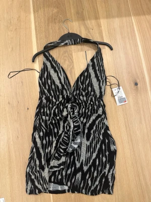 Svart och grå omlottklänning H&M - Snygg kort omlottklänning från H&M i svart och grått med djup v-ringning och knyt framtill. Klänningen har ett zebrainspirerat mönster och halterneck. Perfekt för dig som vill sticka ut med en trendig look. Sitter tajtare än en small, ungefär som en xs då dragkedjan e lite svår att dra upp vid större höfter.