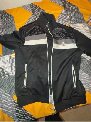 Svart vindjacka från Moncler - Snygg svart vindjacka från Moncler med vit dragkedja, vita detaljer och grått band över bröstet. Jackan har hög krage, två fickor med dragkedja och Moncler-logga på bröstet. Materialmix med glansiga partier upptill ger en modern look.