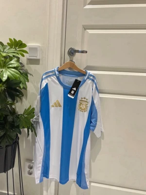 Argentina fotbollströja Adidas - De klassiska blåvita ränderna tillsammans med de exklusiva gulddetaljerna gör detta till en av de snyggaste landslagströjorna. Perfekt för matchdagar, samling eller vardagsbruk.  ✅ Helt ny med tags ✅ Storlek M ✅ Skickas samma dag 📦 ✅ Perfekt inför VM 2026 ✅ Endast 399 kr  🔥 Begränsat pris – först till kvarn!  Argentina, VM 2026, FIFA World Cup 2026, Messi, Lionel Messi, Argentina tröja, Argentina jersey, Adidas, fotbollströja, landslagströja, AFA, världsmästare, football shirt