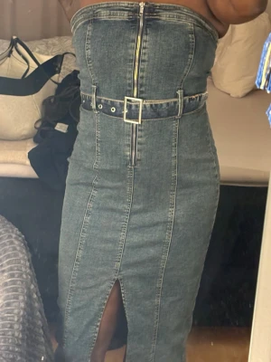 Jeansklänning med bälte och slits - Trendig axelbandslös jeansklänning i mörkblå denim med markerad midja, bälte med silverfärgat spänne och dragkedja framtill. Klänningen har en snygg slits framtill och figurnära passform.