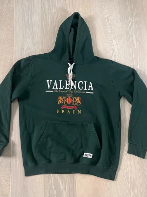 Mörkgrön Valencia hoodie med tryck - Mörkgrön hoodie med stor huva och vitt snöre. Framtill står det 'VALENCIA' och 'SPAIN' i vitt och guld, samt ett färgglatt emblem med lejon. Stor magficka och ribbade muddar. Perfekt för dig som gillar streetwear och vill sticka ut.