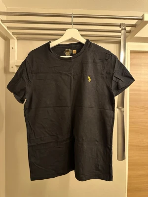 Svart t-shirt från Polo Ralph Lauren - Klassisk svart t-shirt från Polo Ralph Lauren med gul broderad logga på bröstet. Modellen har rund halsringning och korta ärmar. Tillverkad i mjuk bomull och har en slim fit passform som sitter snyggt på kroppen.