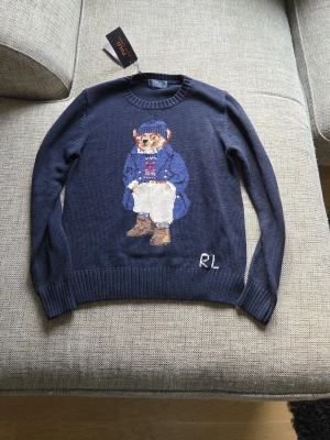 Mörkblå Polo Bear stickad tröja - Oanvänd stickad tröja i mörkblått från Polo Ralph Lauren med ikoniska Polo Bear-motivet framtill. Rund halsringning och ribbade muddar vid ärmslut och nederkant. Tröjan har RL-broderi nedtill och är gjord i mjukt material, perfekt för en klassisk och cool look.