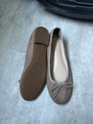 Brun ballerinaskor med rosett - Säljer ett par stilrena bruna ballerinaskor med liten rosett framtill. Skorna har rund tå och ovandel i mockaimitation, samt en bekväm innersula i beige. Yttersulan är i gummi med bra grepp. Perfekta för dig som gillar en enkel och klassisk look.