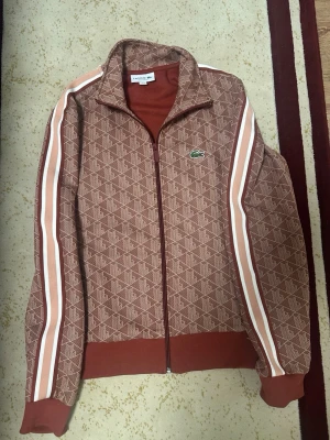 Brun ziptröja från Lacoste - Säljer en brun ziptröja från Lacoste med mönstrad design och vita samt röda ränder längs ärmarna. Tröjan har hög krage, dragkedja framtill och ribbade muddar. Klassisk Lacoste-logga på bröstet. Perfekt för en sportig och trendig look.