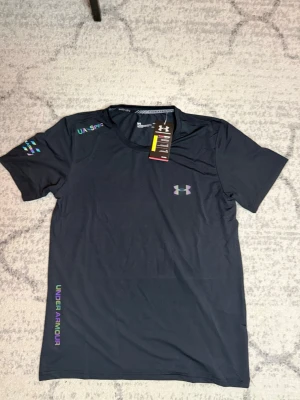 Svart träningströja från Under Armour - Snygg svart funktions-tshirt från Under Armour med regnbågsfärgat logotryck på bröstet och färgglada detaljer på ärmen och sidan. Tillverkad i ett lätt och snabbtorkande material som passar perfekt till träning och sport.