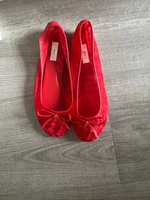 Röda spetsballerinor med rosett från Nelly  - Snygga röda ballerinaskor från Nelly med spetsdetaljer och liten rosett framtill. Skorna har rund tå och är tillverkade i textil med en elegant spetsöverdel. Perfekta för dig som vill sticka ut med färg och stil.
