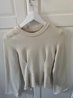 Beige långärmad topp med volangkant - En enkel och stilren beige långärmad topp med rund hals och volangkant nedtill. Toppen har en mjuk struktur och är lätt att matcha till olika outfits. Perfekt för dig som gillar minimalistisk stil.
