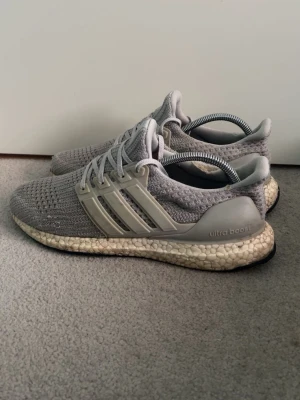 Adidas Ultra Boost  - Säljer ett par grå Adidas Ultra Boost sneakers med stickad ovandel och vita Boost-sulor. Skorna har de klassiska tre ränderna på sidan i beige plast och svart yttersula. Perfekta för dig som gillar stilrena och bekväma sneakers.