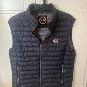 Colmar väst strl 48 - Snygg mörkblå dunväst från Colmar med quiltad design och hög krage. Västen har dragkedja framtill, två fickor med dragkedja och Colmar-logga på bröstet. Perfekt för lager-på-lager och streetstyle. Storlek 48, bra skick.