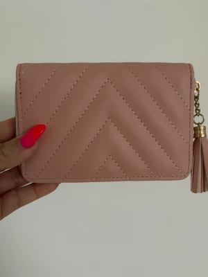 Rosa quiltad plånbok med tofs - Söt rosa plånbok i quiltad design med chevronmönster. Plånboken har dragkedja i guld och en matchande tofs som detalj. Tillverkad i konstläder och har en kompakt, rektangulär form som är lätt att bära med sig.