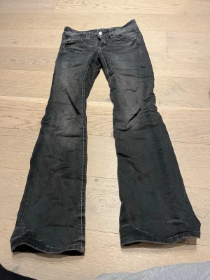 Svarta bootcut jeans med vit söm - Säljer ett par svarta bootcut jeans med vit kontrastsöm och klassisk femficksmodell. Jeansen har låg midja och utsvängda ben, perfekta för en retroinspirerad look. Materialet är denim i bomull. Stängs med knapp och dragkedja framtill.