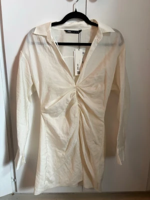 Vit klänning från Zara - Trendig vit kortklänning från Zara med lång ärm. Klänningen har krage och djup v-ringning, vilket ger en modern och stilren look. Tillverkad i ett lätt material som passar perfekt till varmare dagar och student. Aldrig använd men har en liten fläck från ett klistermärke som satt på när jag köpte den, men går säkert bort i tvätten