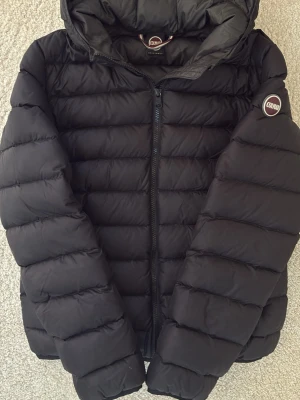 Svart dunjacka från Colmar ungdom - Snygg svart dunjacka från Colmar med huva och dragkedja framtill. Jackan har ett quiltat mönster och Colmar-logga på ärmen. 