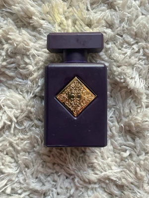 Initio Atomic Rose Eau de Parfum 90ml - Initio Atomic Rose Eau de Parfum i en lyxig mörklila flaska med guldfärgad dekor på framsidan. Flaskan har en fyrkantig form och matchande mörklila lock. Förpackningen är också mörklila med gulddetaljer och text. Innehåller 90 ml.