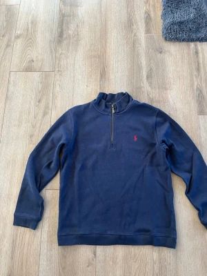 Marinblå half-zip tröja från Polo Ralph Lauren - Klassisk marinblå långärmad tröja från Polo Ralph Lauren med half-zip och ståkrage. Tröjan har det ikoniska röda Polo-logot på bröstet och är tillverkad i mjukt bomullsmaterial. Perfekt för en avslappnad men stilren look.