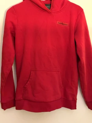 Röd hoodie från Peak Performance - Snygg röd hoodie från Peak Performance med klassisk känguruficka och liten logga på bröstet. Hoodien har huva och ribbade muddar vid ärmslut och nederkant. Perfekt för en avslappnad och sportig stil.