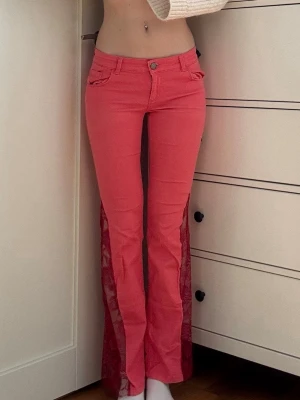 Rosa utsvängda jeans med spetsdetaljer - Säljer ett par unika rosa jeans med utsvängda ben och transparenta spetsinslag längs sidorna. Jeansen har klassisk femficksmodell, knappgylf och cool retrovibe. Perfekta för dig som vill sticka ut och älskar färg och detaljer. Handsydda!