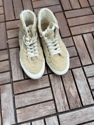 Beige teddy-sneakers från Vans - Unika beige sneakers från Vans med mjukt teddy-material över hela skon. Klassisk hög modell med vita snören och vit sula. Perfekta för dig som vill sticka ut med en mysig och trendig look. Vans-logga på plösen och sidan. Endast använda en gång 