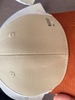 Beige och vit keps från BOSS - Snygg keps från BOSS i beige och vitt med brunt skärm. Framsidan har diskret BOSS-logga och baksidan är i mesh för extra ventilation. Klassisk sexpanel-design och justerbar passform. Perfekt för en avslappnad streetwear-look.