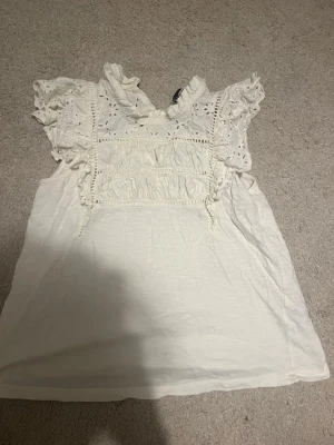 Vit blus med volang och broderi - Supersöt vit blus med volangärmar och broderade detaljer upptill. Blusen har romantiska spetsinslag och en luftig passform, perfekt för varma dagar. Materialet känns lätt och andas bra. Perfekt att styla med jeans eller kjol.